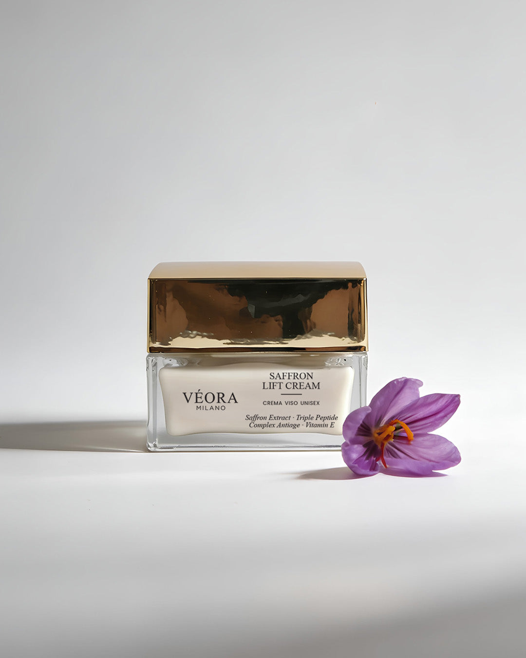 Vasetto di crema Véora allo zafferano con fiore viola su sfondo bianco